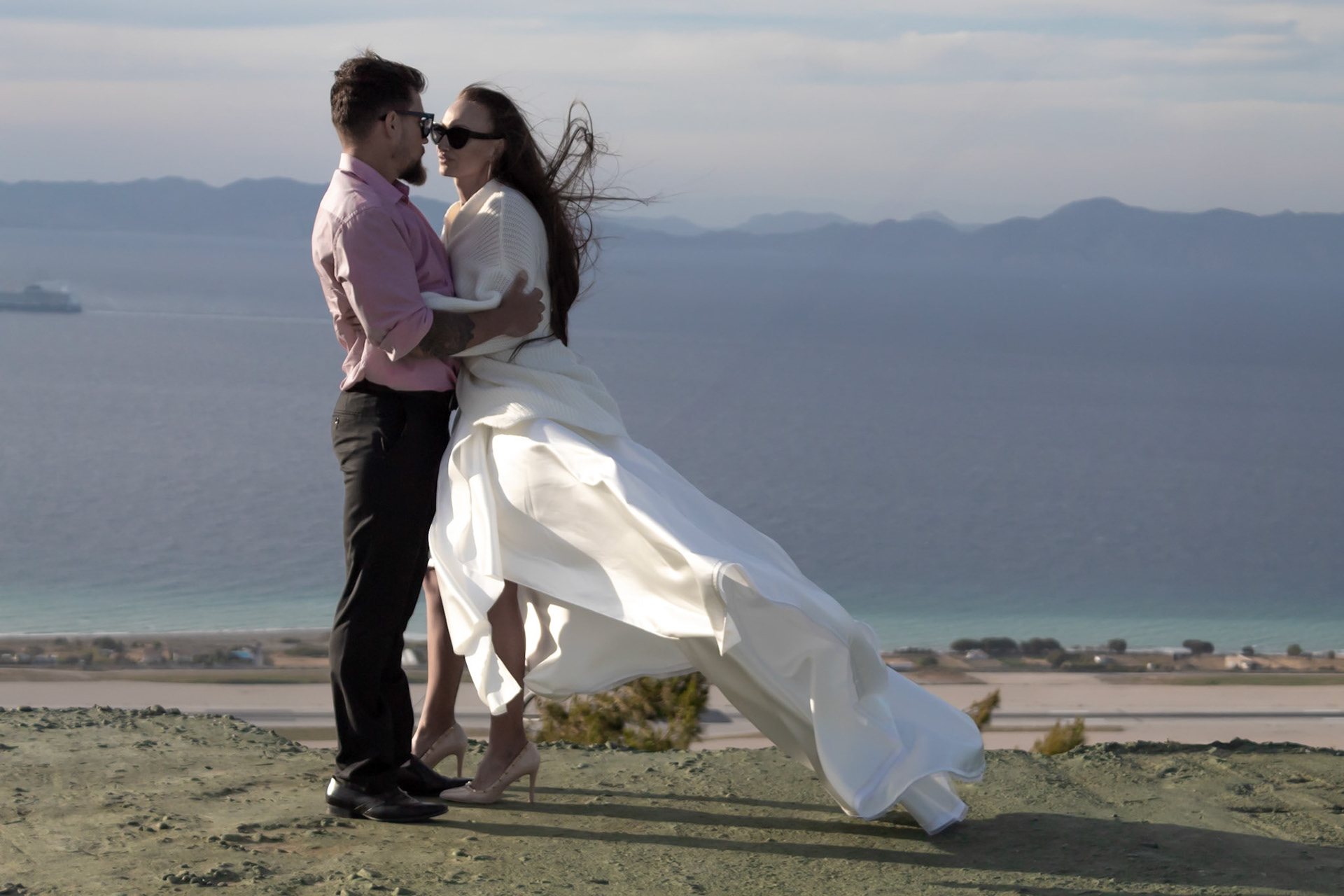 Anna und Aris küssen sich vor dem Meer, während der Wind das Brautkleid bewegt